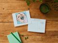 A flat-lay of a custom pet portrait postcard and a matching postcard with a 'Treats Please!' stamp design, arranged on a rustic wooden table with a green notebook, pencil, and a glass of water.