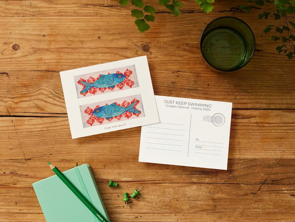 A styled flat-lay of a postcard set on a rustic wooden table. The front of the postcard features two blue fish swimming over a red patterned background, and the back displays the text 'JUST KEEP SWIMMING' with a postmark seal.