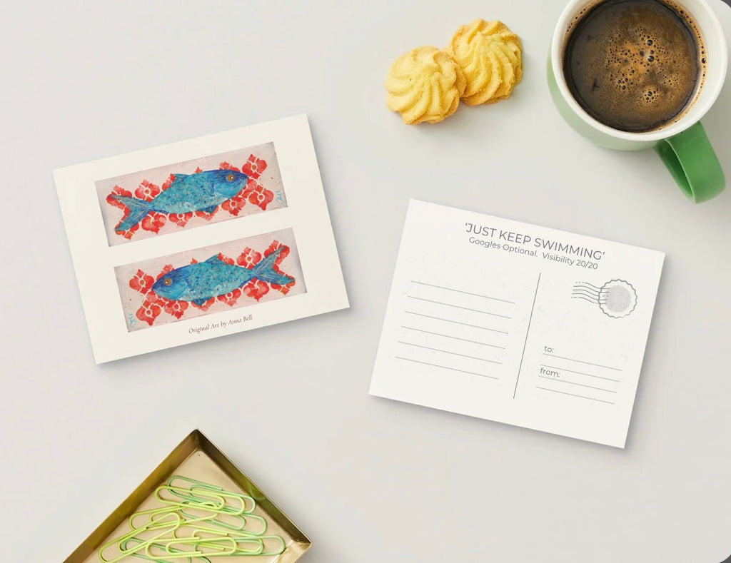 A postcard set displayed on a light gray surface, featuring a front card with two blue fish illustrations surrounded by red hearts, and a back card with the text 'JUST KEEP SWIMMING' and 'cooler than yesterday, visibility 2020'. A green coffee mug, two yellow cookies, and a tray of green paperclips are arranged around it as styled props.