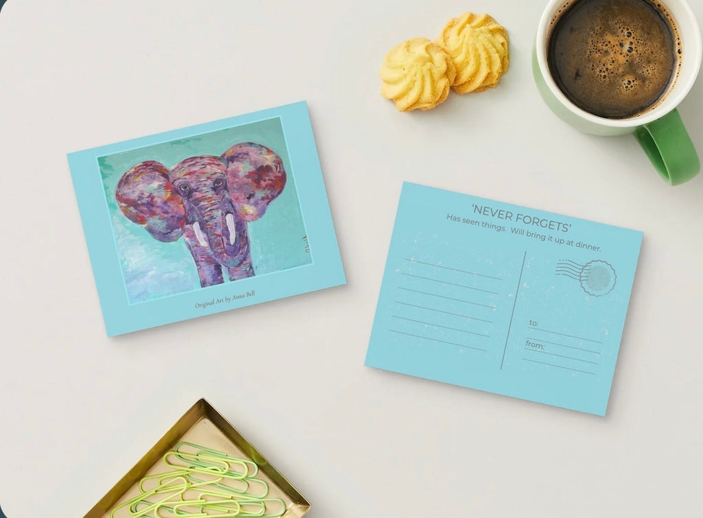 A styled flat-lay of a postcard set on a white surface, featuring a front card with a colorful elephant illustration and a back card with writing lines and the text 'NEVER FORGETS'. Yellow cookies, a green mug of coffee, and paper clips are arranged around them.