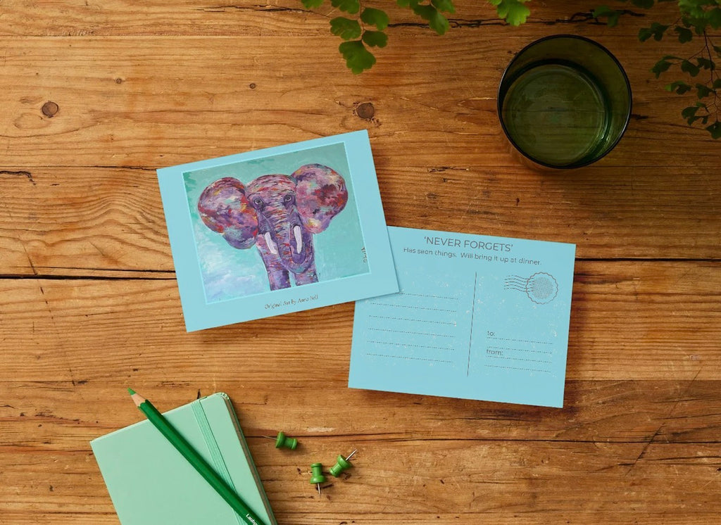 A postcard set displayed on a rustic wooden table, featuring a front card with a colorful elephant illustration and a back card with writing sections and a circular stamp area. A mint green notebook, green pencil, and a dark green glass cup are arranged nearby as props.