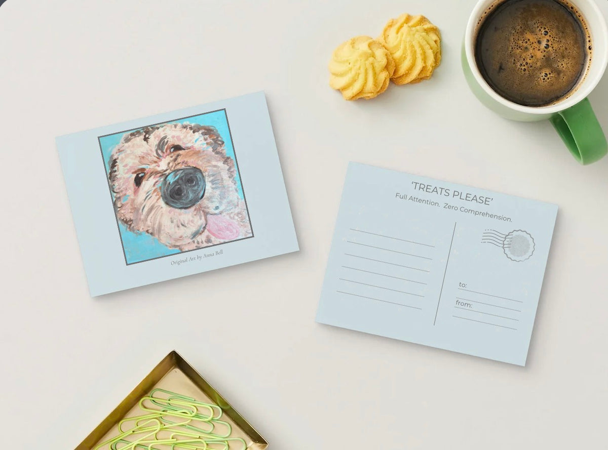 A flat-lay of a dog-themed postcard set on a white surface, featuring a front card with a colorful sketch of a dog and a back card with writing lines and a postage stamp illustration. A green coffee mug, two yellow cookies, and paperclips are styled around it.