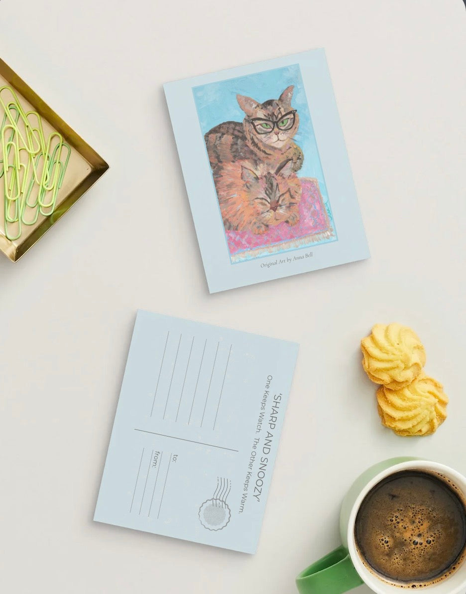 A styled flat-lay featuring a postcard with a colorful illustration of a cat and another postcard with the text 'SHARP AND SNOOZY One Sharp Watch. The Other Sleeps Warm.