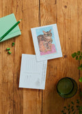 A flat-lay of two 'Tharp and Snoopy' art postcards on a rustic wooden table, surrounded by a green notebook, pencil, binder clips, and a glass of water.