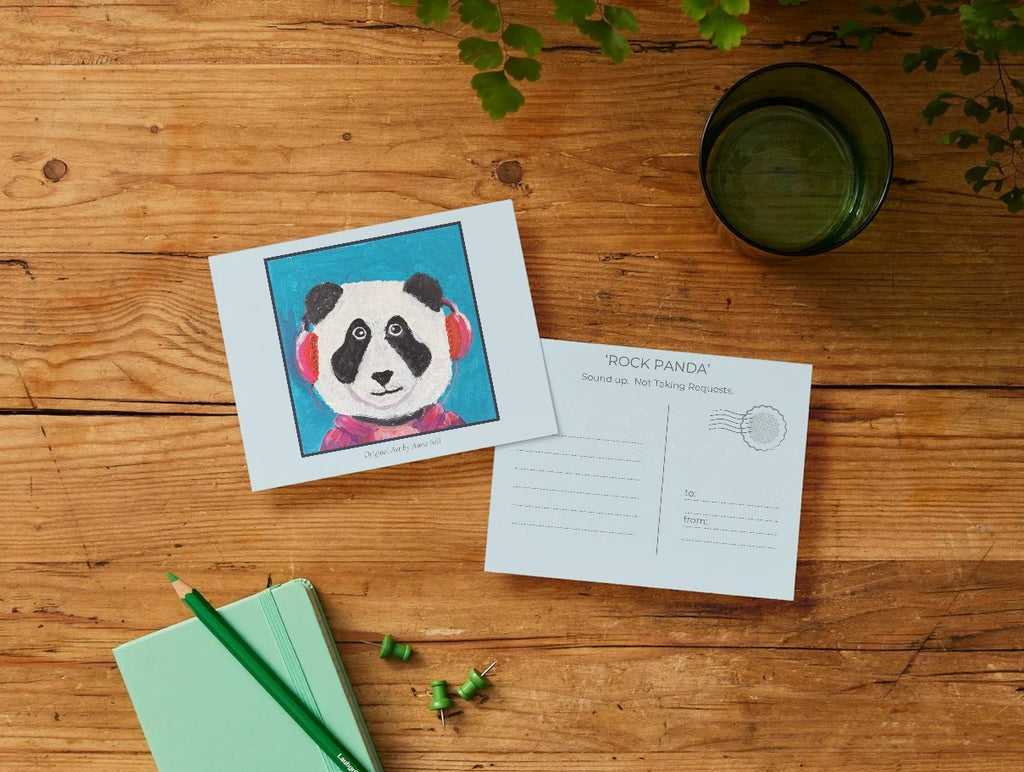 A printed card or postcard featuring a colorful panda illustration lies on a wooden tabletop next to the back of another postcard with printed writing areas and a stamp box. A mint notebook with a green pencil, loose green paper clips, and a dark green drinking glass are also visible in a styled flat-lay arrangement.