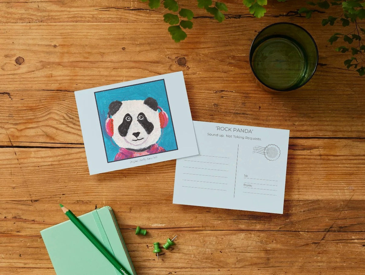 A printed card or postcard featuring a colorful panda illustration lies on a wooden tabletop next to the back of another postcard with printed writing areas and a stamp box. A mint notebook with a green pencil, loose green paper clips, and a dark green drinking glass are also visible in a styled flat-lay arrangement.