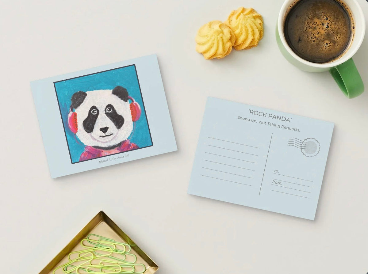 A postcard set featuring a colorful illustration of a panda wearing pink headphones on one card and a blank back with writing lines and a postage stamp design on another. The cards are arranged on a light surface with a green coffee mug, cookies, and paper clips nearby.