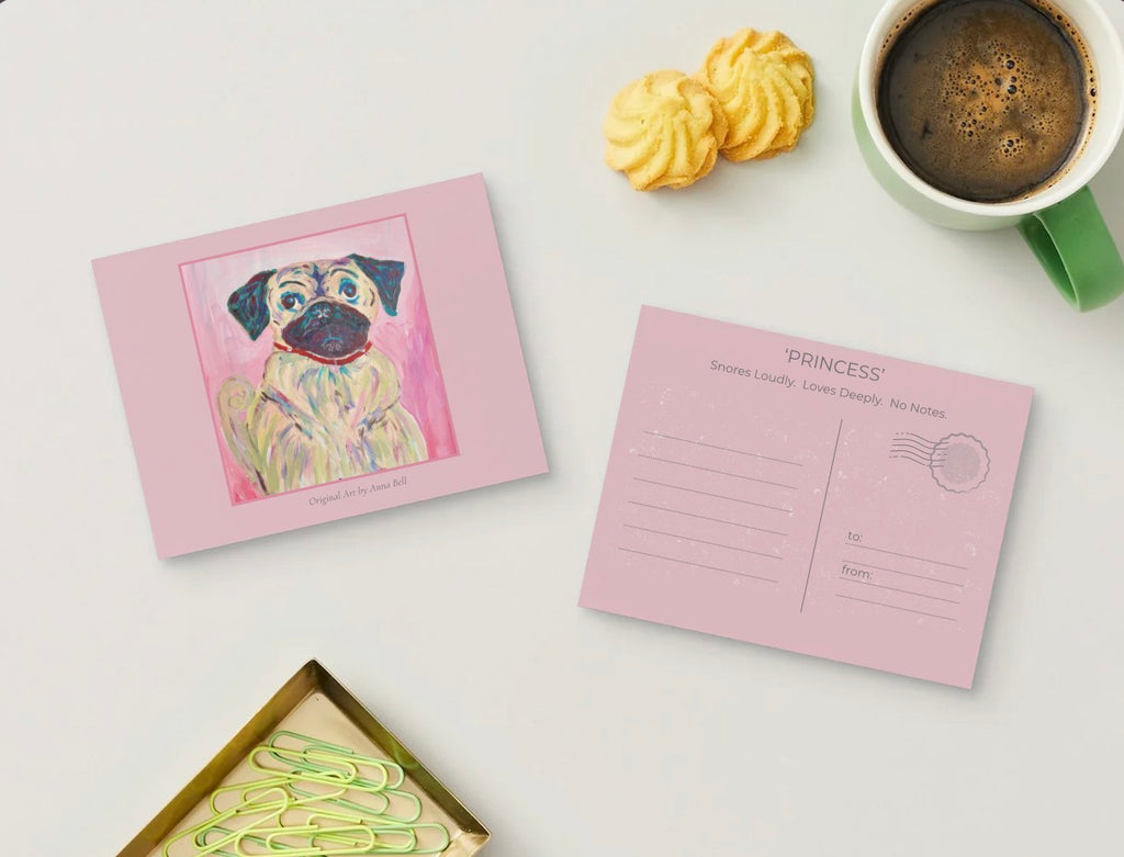 A pink postcard set displayed on a light gray surface, featuring one front side with a colorful painted pug illustration and one back side with 'PRINCESS' lettering and postmark lines. Yellow cookies, a green mug of coffee, and paperclips are arranged around it as props.