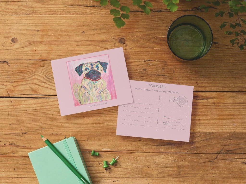 A pink postcard set displayed on a rustic wooden table, featuring a front card with a colorful illustration of a pug and a back card with printed writing and stamp fields. Styled flat-lay props include a mint green notebook with green pencils, a dark green glass, and green leaves.