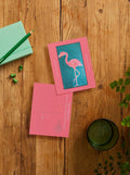 A styled flat-lay of two pink postcards on a rustic wooden table. One postcard features a vibrant pink flamingo illustration on a teal background, while the other is a pink postcard with 'PALM ROYALE' printed on it.