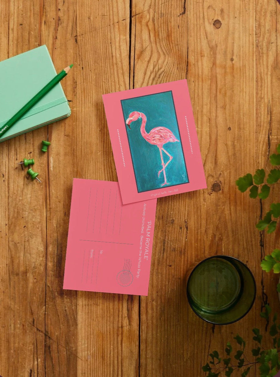 A styled flat-lay of two pink postcards on a rustic wooden table. One postcard features a vibrant pink flamingo illustration on a teal background, while the other is a pink postcard with 'PALM ROYALE' printed on it.