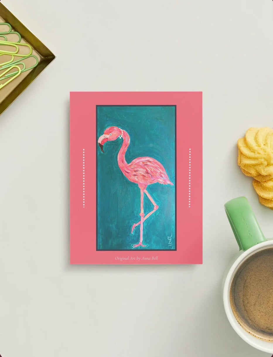 A pink art print featuring a painted pink flamingo on a teal background, styled on a light gray surface with a green mug, cookies, and a decorative tray nearby.