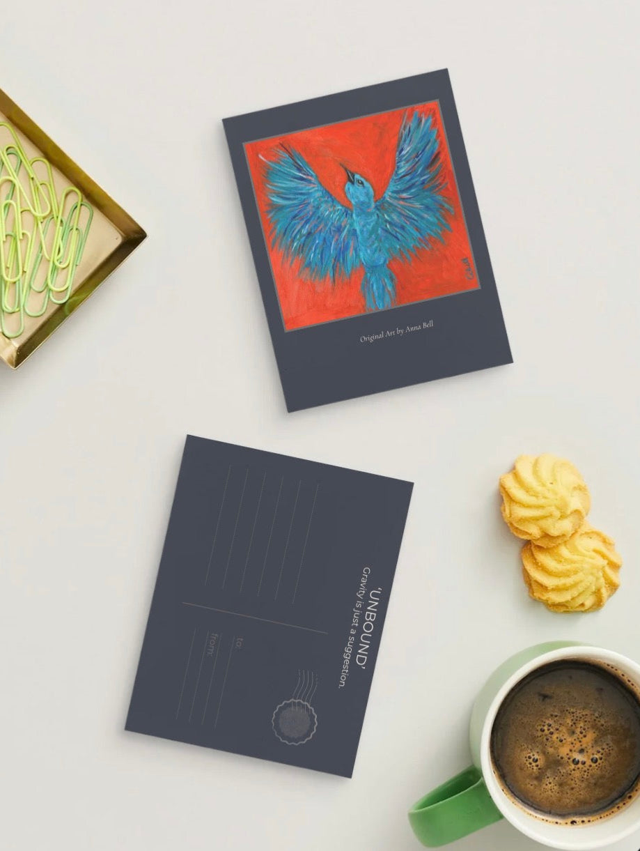 A styled flat-lay of two dark gray postcards on a light gray surface, surrounded by cookies, a cup of coffee, and paperclips. One postcard features a vibrant illustration of a blue bird on a red background, and the other is blank with the title 'UNBOUND'.
