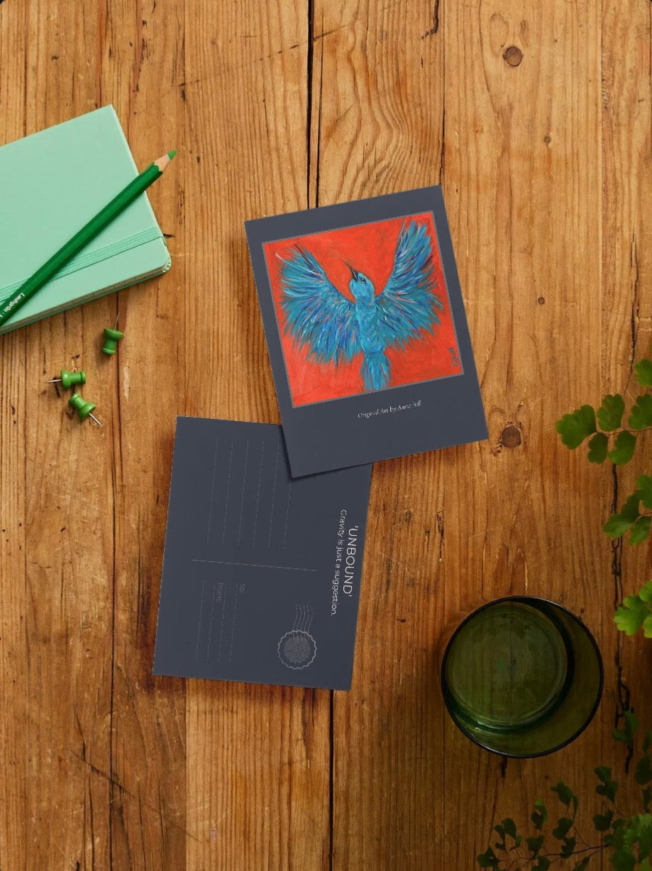 A postcard set displayed on a rustic wooden tabletop, featuring a front card with a vibrant blue bird illustration on a red background and a back card with 'UNBOUND' printed on it. Styled flat-lay props include a green notebook, pencil, and a glass of water.